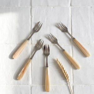May include: A collection of five small forks with light-colored handles and silver tines, arranged on a white tiled surface. A single stalk of wheat is placed near the forks, suggesting a rustic or farmhouse aesthetic.