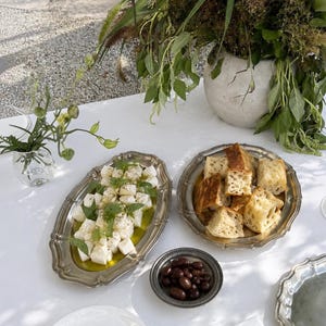 Peut inclure: Un plateau ovale en argent contient des cubes de fromage blanc arrosés d'huile d'olive et garnis d'herbes vertes. Un plateau rond en argent contient du pain en cubes. Un petit bol en argent contient des olives noires. Assiettes et nappe blanches.
