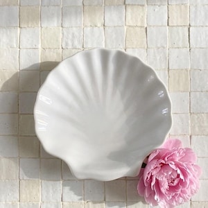 May include: A white, shell-shaped ceramic dish with a scalloped edge. A pink peony flower rests beside the dish on a tiled surface. The dish is a decorative item, suitable for serving or display.