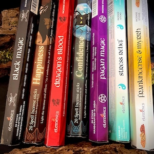 May include: A selection of incense stick boxes, with labels like "Black Magic" and "Dragon's Blood." The boxes are in various colors, including black, red, and purple. They are displayed on a dark, textured surface, with text in English.