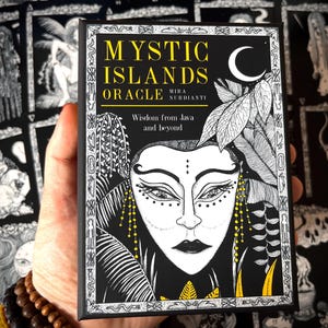 May include: A black and white Mystic Islands Oracle card deck. The cover features a detailed illustration of a face with a crescent moon and foliage. Text includes "Mystic Islands Oracle" and "Wisdom from Java and beyond".