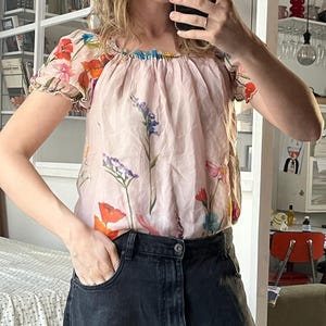 Puede incluir: Una blusa de seda rosa con estampado floral. La blusa tiene un escote fruncido y mangas cortas. Las mangas tienen un detalle de volantes. La blusa est&aacute; metida en un par de pantalones vaqueros negros.