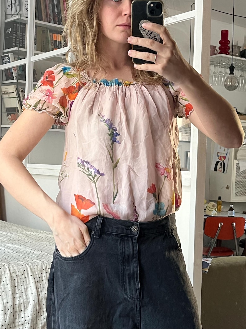 Puede incluir: Una blusa de seda rosa con estampado floral. La blusa tiene un escote fruncido y mangas cortas. Las mangas tienen un detalle de volantes. La blusa est&aacute; metida en un par de pantalones vaqueros negros.
