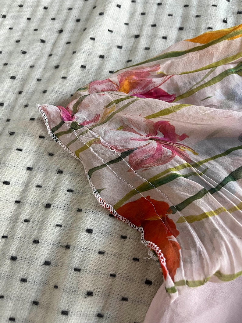 Puede incluir: Primer plano de una tela rosa transparente con estampado floral. La tela tiene un ribete blanco y est&aacute; doblada sobre s&iacute; misma. El estampado floral presenta colores rosa, naranja y verde.