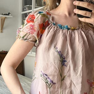 Puede incluir: Una blusa rosa con estampado floral, cuello fruncido y mangas cortas abullonadas. La blusa presenta una variedad de dise&ntilde;os florales, incluyendo amapolas, flores silvestres y otras plantas.