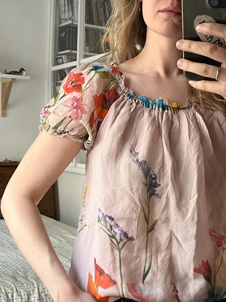 Puede incluir: Una blusa rosa con estampado floral, cuello fruncido y mangas cortas abullonadas. La blusa presenta una variedad de dise&ntilde;os florales, incluyendo amapolas, flores silvestres y otras plantas.