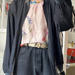 Puede incluir: Una chaqueta bomber negra con cierre de cremallera y una blusa de estampado floral debajo. La chaqueta tiene cuello y pu&ntilde;os de canal&eacute;. La blusa tiene un escote con volantes y un estampado floral con flores rosas, azules y rojas. La persona lleva pantalones vaqueros negros y un cintur&oacute;n de piel de serpiente.