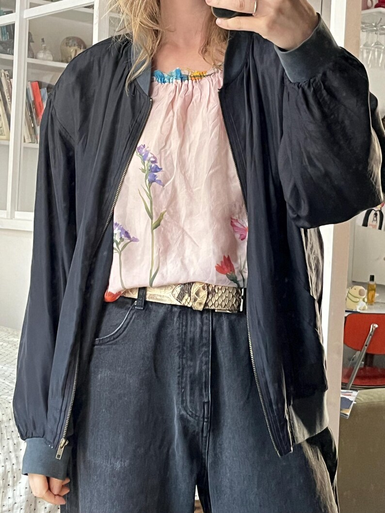 Puede incluir: Una chaqueta bomber negra con cierre de cremallera y una blusa de estampado floral debajo. La chaqueta tiene cuello y pu&ntilde;os de canal&eacute;. La blusa tiene un escote con volantes y un estampado floral con flores rosas, azules y rojas. La persona lleva pantalones vaqueros negros y un cintur&oacute;n de piel de serpiente.