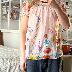 Puede incluir: Una blusa rosa con estampado floral con una variedad de flores de colores. La blusa tiene un escote fruncido y mangas cortas.