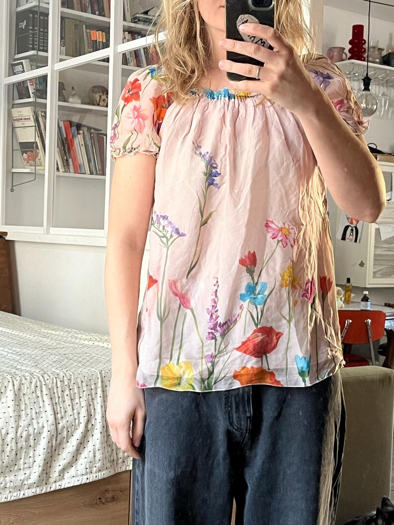 Puede incluir: Una blusa rosa con estampado floral con una variedad de flores de colores. La blusa tiene un escote fruncido y mangas cortas.