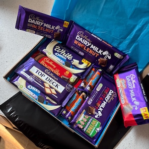 Puede incluir: Una caja de regalo llena de varias barras de chocolate Cadbury. La selección incluye Dairy Milk con Hazelnut, Whole Nut, White Oreo, Bournville, Top Deck, Fudge y Fruit & Nut. Las barras están en su embalaje original, con marca púrpura y blanca.