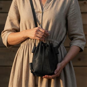 May include: A black leather bucket bag with a drawstring closure and a long, adjustable strap. The bag is held by a person wearing a beige dress. The background is a wooden wall.