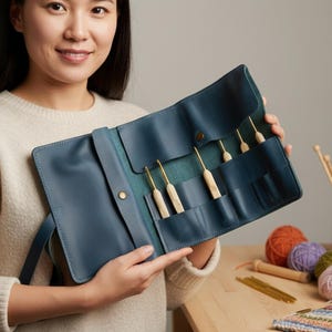 May include: A woman holds an open, blue leather crochet hook case. The case contains several crochet hooks with wooden handles and gold-colored metal tips. The case is open, revealing the hooks, and is displayed against a neutral background with yarn and knitting needles.