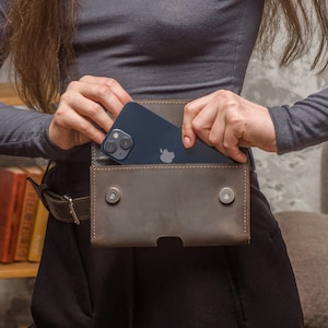 May include: A dark brown leather belt pouch featuring a rectangular flap with two silver-toned magnetic snap closures. The pouch is secured to a matching leather belt and contains a dark blue smartphone with a visible dual-camera system.