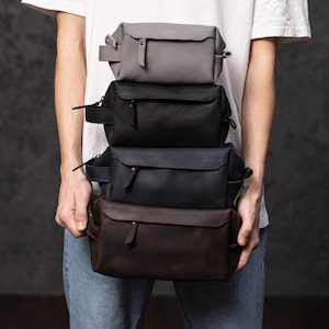 May include: Four leather wash bags in grey, black, navy, and brown are stacked. Each bag has a zip closure and a small handle. The rectangular bags appear to be made of smooth leather. The bags are held by a person.