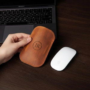 Könnte beinhalten: Eine braune Leder-Maus-Hülle mit den Initialen "JK" in einem Kreis, neben einer weißen Computermaus. Im Hintergrund ist ein Laptop zu sehen. Die Hülle wird von einer Hand gehalten. Die Artikel befinden sich auf einer dunklen Holzoberfläche.