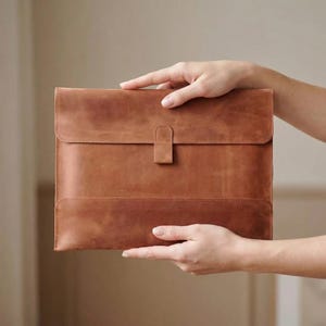Puede incluir: Una funda de cuero marrón para portátil con cierre de solapa y lengüeta. La funda está sujeta por dos manos. El cuero tiene un aspecto natural y texturizado.