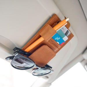 May include: A tan leather car visor organizer featuring a central pen loop, multiple slots for credit cards, and a sturdy strap for securing sunglasses. The organizer is attached to a light-colored vehicle sun visor, holding an orange pen and cards.