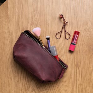May include: A burgundy leather makeup bag with a black zipper, containing makeup brushes and a lipstick. A rose gold eyelash curler and a pink lipstick are next to the bag. The items are on a light wood surface.