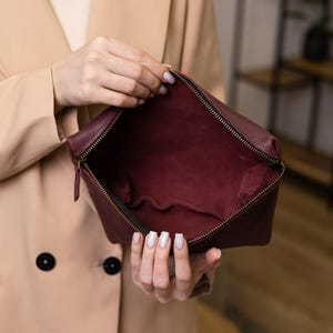 May include: A burgundy leather pouch with a zippered closure is held open, revealing a matching interior. The pouch has a rectangular shape with rounded corners. The zipper is a dark metallic color. The background is blurred.
