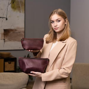 May include: Two burgundy leather fanny packs with zippered closures. The bags are held up by a person wearing a tan blazer. The bags are different sizes, with a rectangular shape.