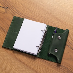 Könnte beinhalten: Offener grüner Lederordner mit Stifthalter und einem Stapel weißem Papier. Der Ordner hat einen Drei-Ring-Mechanismus und ist auf einer Holzoberfläche geöffnet. Ein lila Stift befindet sich im Stifthalter.