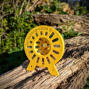 Peut inclure: Un objet circulaire jaune vif avec des formes découpées et le texte "la-bonne-com.fr" sur le bord. L'objet a deux petits pieds et repose sur un morceau de bois vieilli. L'arrière-plan est flou avec du feuillage vert.