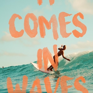 May include: A woman surfs a wave in the ocean. The sky is blue and the water is turquoise. The words "IT COMES IN WAVES" are written in orange paint across the image.
