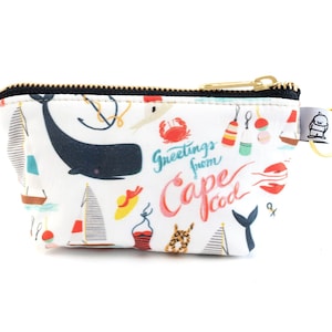 May include: A white zippered pouch with a colorful print of nautical themed illustrations, including a whale, boats, and a lobster. The text "Greetings from Cape Cod" is printed on the pouch.