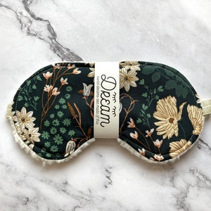 May include: A dark green sleep mask with a floral pattern in cream and brown. The mask has a soft, white, fluffy trim and a cream-coloured elastic strap. The text "Dream" and "sweet dreams are made of this" is printed on a white label.