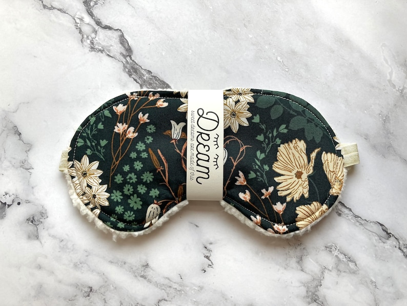 May include: A dark green sleep mask with a floral pattern in cream and brown. The mask has a soft, white, fluffy trim and a cream-coloured elastic strap. The text "Dream" and "sweet dreams are made of this" is printed on a white label.