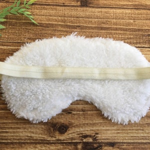 May include: A white, fluffy sleep mask with a cream elastic strap. The mask is made of a soft, textured material, designed to block out light. The background is a wooden surface.
