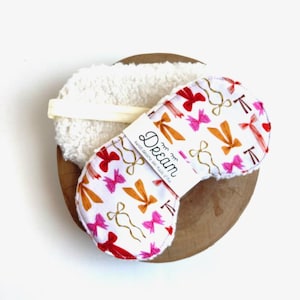 May include: A white sleep mask with a pattern of red, pink, and gold bows, and the word "Dream" printed on a white label. The mask is paired with a white, fluffy face pad, all presented on a wooden surface.