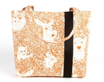 May include: Tote bag with a floral and animal print in orange and white. The bag features illustrations of a fox, deer, rabbit, and llama. A vertical black stripe runs down the side, and the handles are brown.