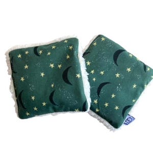 May include: Two green fabric hand warmers with a white sherpa trim. The fabric has a pattern of yellow stars and black crescent moons.
