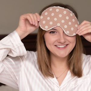 May include: A beige sleep mask with white polka dots and a white trim. The sleep mask is being held up by a person wearing a pink and white striped pajama top. The background is a blurred bedroom setting.