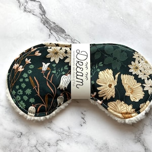 May include: A dark green sleep mask featuring a cream and tan floral design. The mask has a soft, white, fluffy trim and a band with the word "Dream" printed on it. The text "sweet dreams are made of this" is also printed on the band.