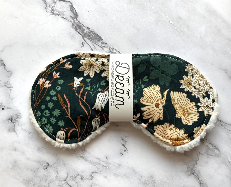 May include: A dark green sleep mask featuring a cream and tan floral design. The mask has a soft, white, fluffy trim and a band with the word "Dream" printed on it. The text "sweet dreams are made of this" is also printed on the band.