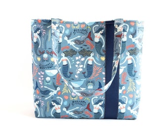 May include: A blue tote bag with a pattern of mermaids, whales, and seaweed. The bag has navy blue straps.