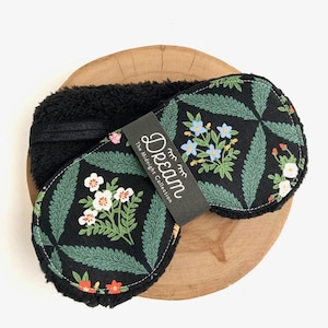May include: Black sleep mask with a floral pattern in green, white, and red on a black background. The mask has a soft, black, plush lining and a black elastic band. A tag reads "Dream The Midnight Collection."