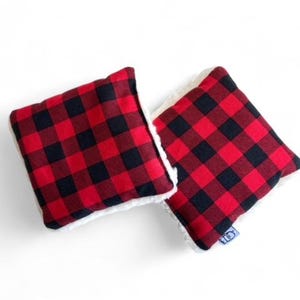 May include: Two red and black buffalo plaid fabric squares with white sherpa trim. The squares are sewn together at the edges.