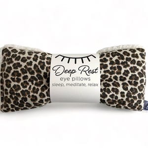 May include: A Deep Rest eye pillow with a leopard print pattern. The pillow has a white, fluffy trim on the ends and a white band with the text "Deep Rest eye pillows sleep, meditate, relax". A small tag with a cartoon character is attached.