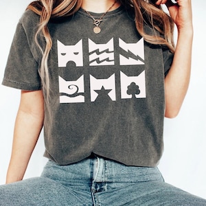 May include: A charcoal gray short-sleeve t-shirt featuring a white graphic print of six square icons arranged in two rows. Each icon depicts a stylized cat head silhouette containing different symbols including waves, a star, a tree, and lightning.