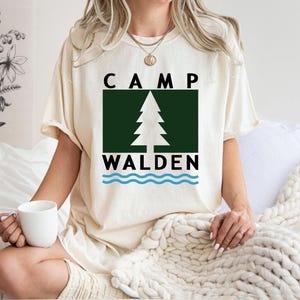 Può includere: Una t-shirt color crema con una stampa grafica con il testo CAMP WALDEN in lettere nere. Il design include un blocco rettangolare verde scuro contenente la sagoma di un pino bianco, posizionato sopra tre linee blu ondulate.