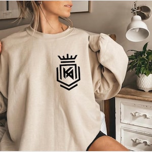 May include: A beige crewneck sweatshirt featuring a black graphic print on the left chest. The design consists of a stylized crown positioned above a geometric shield emblem containing the letters KA in a bold, angular, modern font.