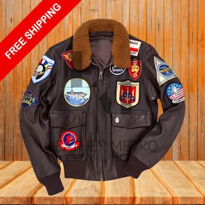 May include: A brown leather bomber jacket with a shearling collar and multiple patches. The patches include a ship, a trident, a seabee logo, a plane, and a tomcat logo. The text on the patches includes "SEABEES", "U.S.C. WILLIAM P. STANDLEY", "01012", and "TOMCAT".