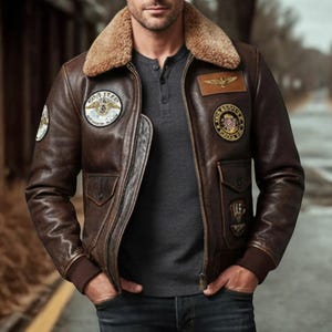 May include: A brown leather jacket with a tan shearling collar. The jacket features multiple patches, including a circular patch on the left sleeve and a rectangular patch on the chest. It has two front pockets and a zipper closure.