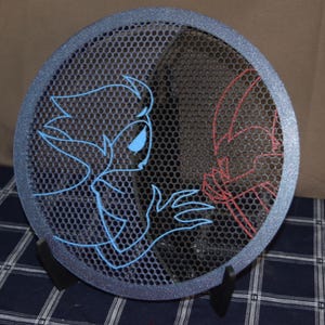 May include: A round, dark blue speaker cover with a mesh design. Outlined in blue and red are cartoon figures. The rim of the cover has a glittery finish. The speaker cover is propped up on a black stand.