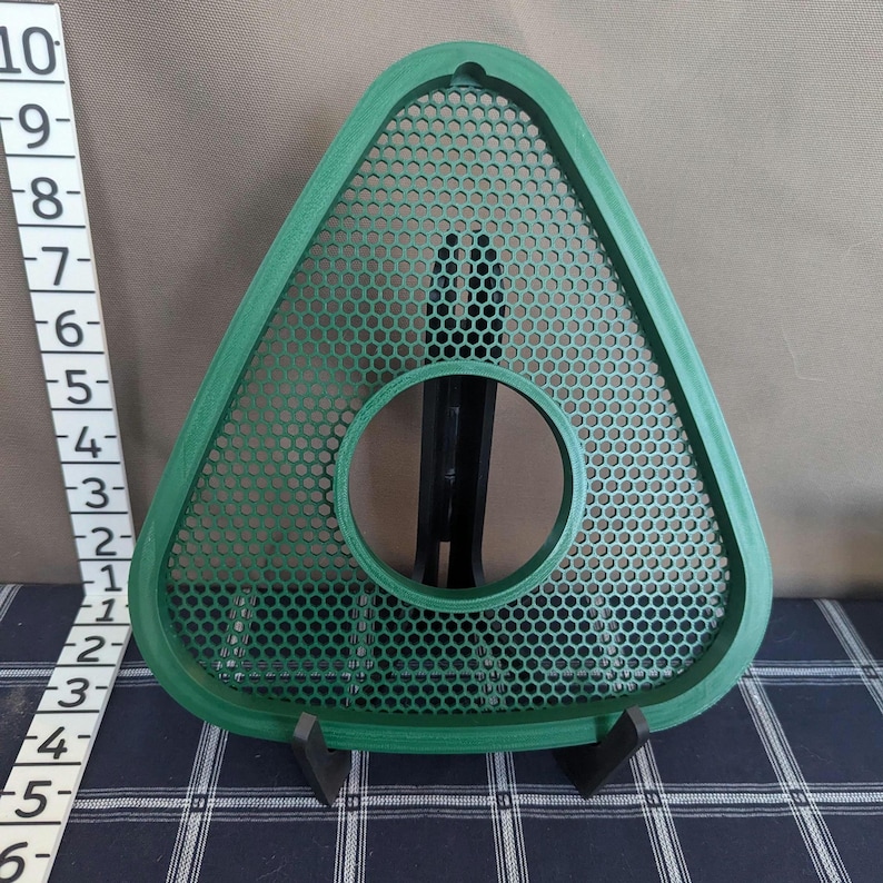 May include: A green, triangular object with a perforated surface and a circular opening. The object is approximately 9 inches tall and is supported by a black stand. The design features a honeycomb pattern.