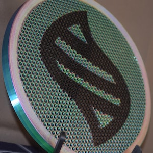 Op de afbeelding: Cirkelvormig, iriserend metalen embleem met een honingraatpatroon en een donkerbruine gestileerde letter "S" in het midden. De rand heeft een regenboogglans, met een blauwe en roze gradiënt. Waarschijnlijk een decoratief item of verzamelobject.